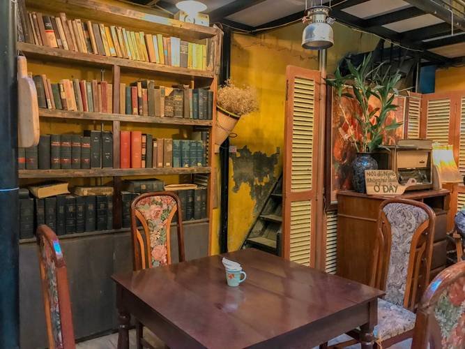 cafe-sach-ha-noi-13-1024x768-1 Một góc quán cà phê ấm cúng với bàn ghế gỗ, kệ sách đầy sách và trang trí cây xanh.