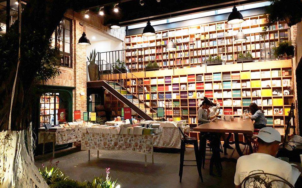 cafe-sach-ha-noi-2 Quán cà phê sách ấm cúng với giá sách lớn, bàn ghế gỗ, ánh sáng dịu và một số người đang ngồi đọc.