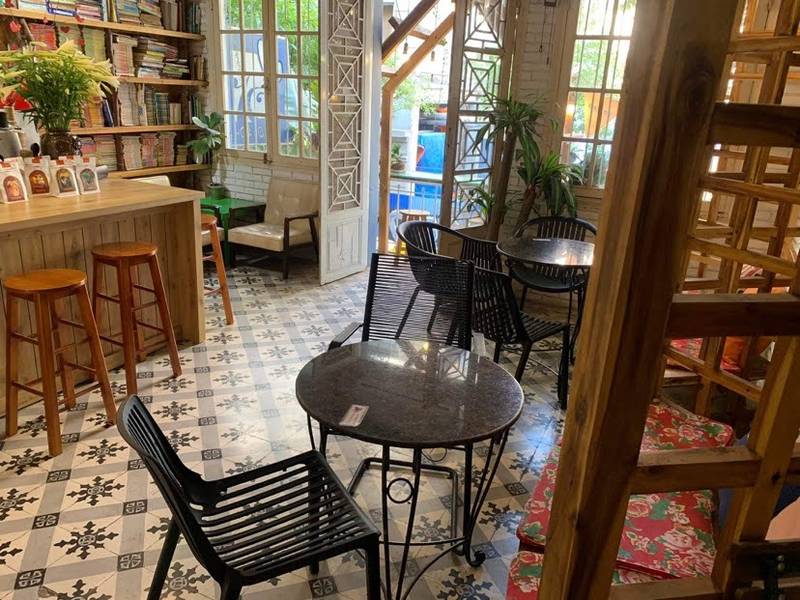 cafe-sach-ha-noi-3 Quán cà phê ấm cúng với bàn ghế nhỏ, sàn gạch hoa, kệ sách và cửa sổ nhìn ra không gian xanh bên ngoài.