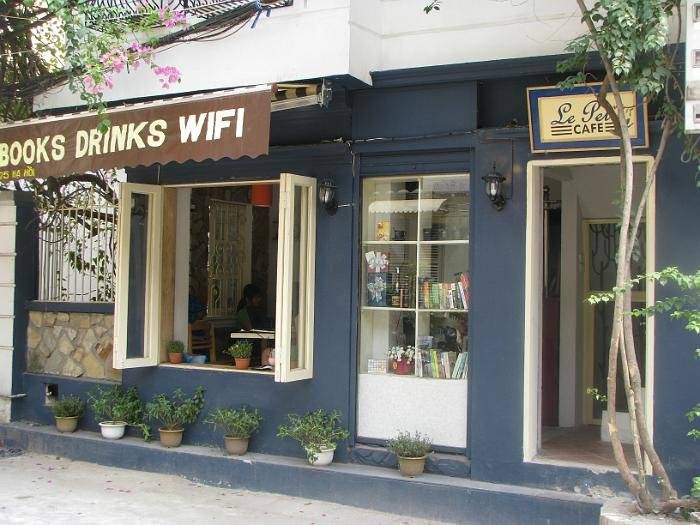 cafe-sach-ha-noi-6 Mặt tiền quán cà phê nhỏ, có biển hiệu với dòng chữ "BOOKS DRINKS WIFI" và "Le Petit Café", trang trí cây xanh.