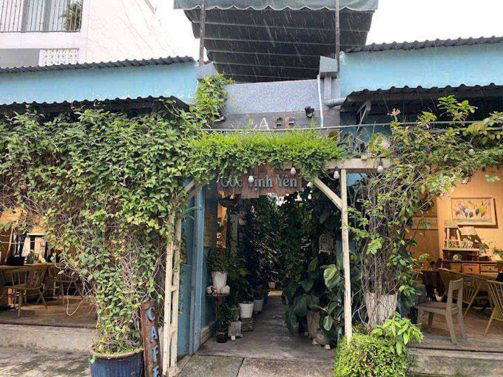Khám Phá Top 10 Quán Cafe Sân Vườn Đẹp và Thư Giãn Tại Thủ Đức 4 Lối vào quán cà phê với cây xanh bao phủ, bên trong có bàn ghế và trang trí tranh ảnh.