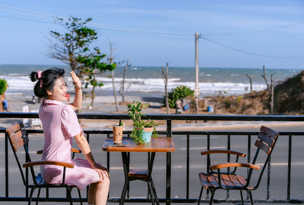 Khám Phá 12+ Quán Cafe Ngắm Hoàng Hôn Tuyệt Đẹp Tại Vũng Tàu 11 Người ngồi trên ban công nhìn ra biển, cạnh bàn có cây xanh và đồ uống, trời nắng đẹp.