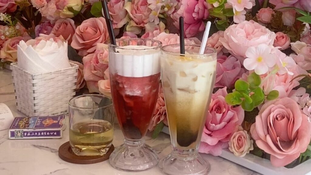 Hai ly nước uống tr&ecirc;n b&agrave;n cạnh b&oacute; hoa hồng, với khăn giấy trong giỏ v&agrave; một ly tr&agrave; nhỏ b&ecirc;n cạnh.