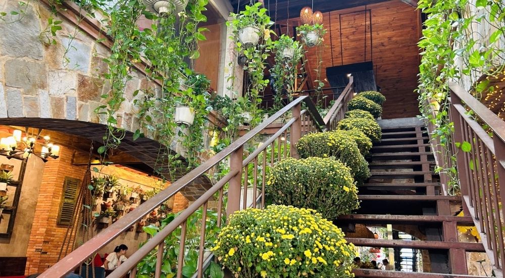 Khám Phá 10 Quán Cafe Gò Vấp Với Góc "Hidden Gem" Độc Đáo 6 Cầu thang sắt trang trí hoa và cây xanh trong quán cà phê, ánh sáng ấm áp và tường gạch tạo không gian thoải mái.