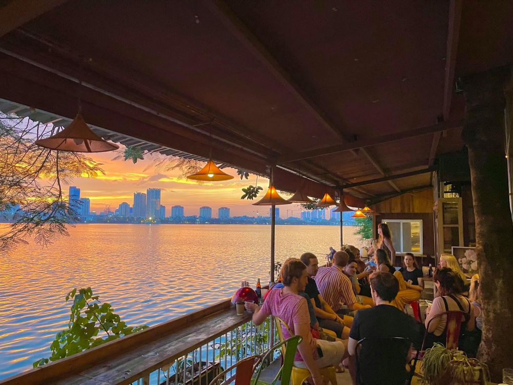 Khám Phá Top 10 Quán Cafe Hồ Tây View Đẹp Ngắm Hoàng Hôn Lãng Mạn 5 Quán cà phê bên hồ với nhóm người trò chuyện dưới ánh đèn vàng, trời hoàng hôn và cảnh thành phố ở xa.