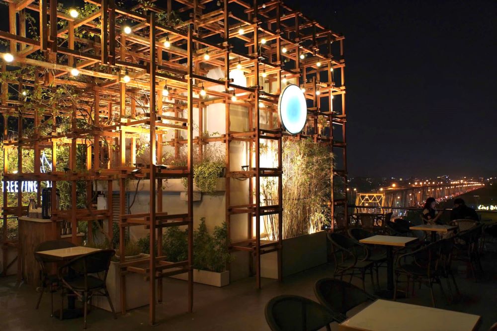 Khám Phá 10+ Quán Cafe Đẹp Nhất Long Biên: Thỏa Sức “Sống Ảo”, Thưởng Thức Đồ Uống Ngon 10 Quán cà phê ngoài trời về đêm, với giàn đèn và trang trí cây xanh, nhìn ra cầu sáng đèn.