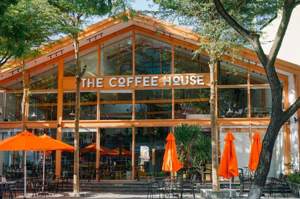 Khám Phá 10+ Quán Cafe Đẹp Nhất Long Biên: Thỏa Sức “Sống Ảo”, Thưởng Thức Đồ Uống Ngon 5 Mặt tiền quán cà phê với mái kính, ô che màu cam và cây xanh xung quanh, tạo không gian thoáng đãng.