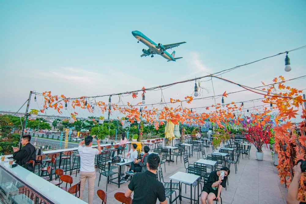 Top 11+ Quán Cafe Đẹp Tại Đà Nẵng Không Thể Bỏ Qua Năm 2025 24 Quán cà phê ngoài trời với máy bay bay qua, xung quanh là trang trí lá cây mùa thu và bàn ghế trống.