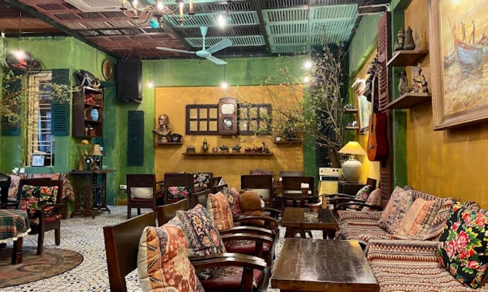 Khám Phá Top 6 Quán Cà Phê Trịnh Hấp Dẫn Tại Cầu Giấy 11 Quán cà phê với nội thất cổ điển, có bàn ghế gỗ, gối hoa văn và trang trí cây xanh, tạo không gian ấm cúng.