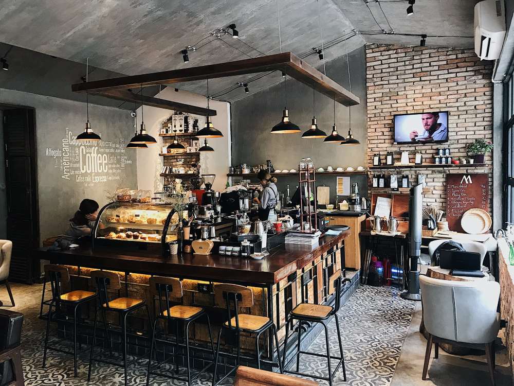 Qu&aacute;n c&agrave; ph&ecirc; với quầy bar gỗ, đ&egrave;n treo, v&agrave; tường gạch, c&oacute; người đang phục vụ ph&iacute;a sau quầy.