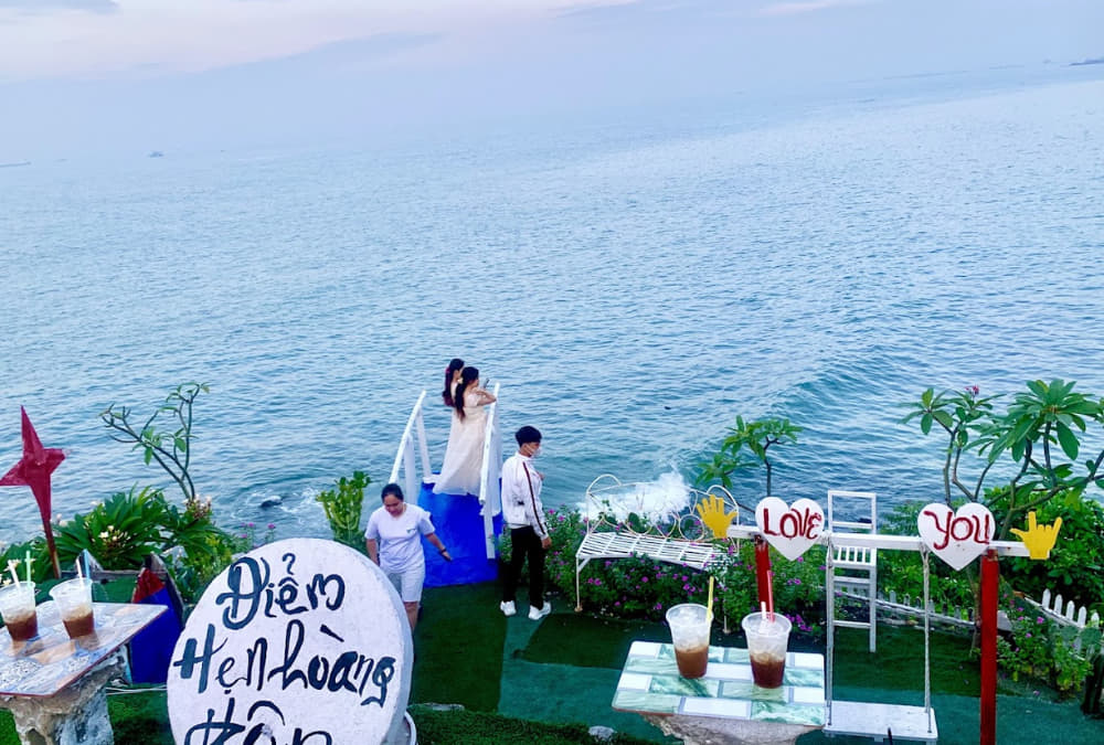 Khám Phá 12+ Quán Cafe Ngắm Hoàng Hôn Tuyệt Đẹp Tại Vũng Tàu 12 Khung cảnh biển với khu vực ngắm cảnh, vài người đứng gần biển, cảnh vật trang trí với bảng hiệu và cây xanh.