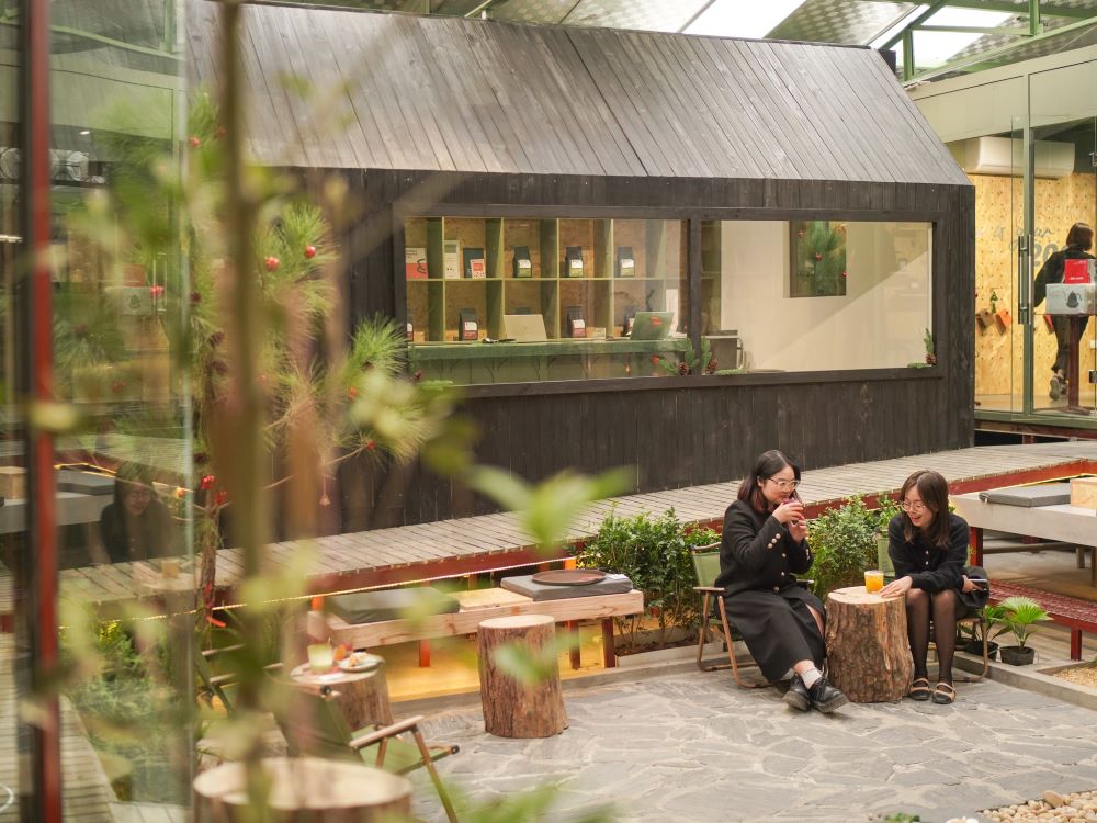 Khám Phá 10+ Quán Cafe Đẹp Nhất Long Biên: Thỏa Sức “Sống Ảo”, Thưởng Thức Đồ Uống Ngon 17 Hai người ngồi trò chuyện trong quán cà phê ngoài trời, phía sau là ngôi nhà gỗ nhỏ và cửa sổ trưng bày.