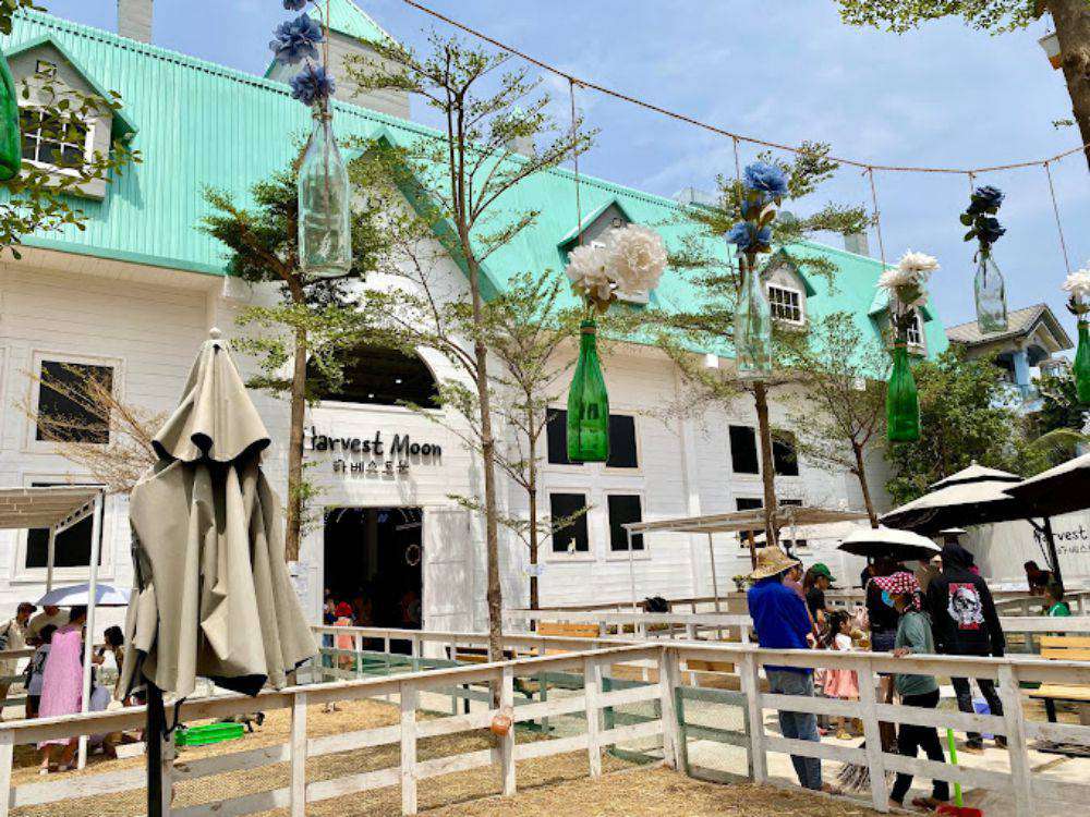 Khám Phá Top 10 Quán Cafe Sân Vườn Đẹp và Thư Giãn Tại Thủ Đức 13 Tòa nhà màu trắng với mái xanh, treo chai thủy tinh trang trí, có nhiều người tham quan và cây xanh xung quanh.
