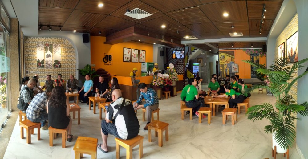 Khám Phá 10+ Quán Cafe Đẹp Nhất Long Biên: Thỏa Sức “Sống Ảo”, Thưởng Thức Đồ Uống Ngon 8 Quán cà phê với nhiều người ngồi trò chuyện trên ghế gỗ, tường trang trí hoa văn và cây xanh xung quanh.