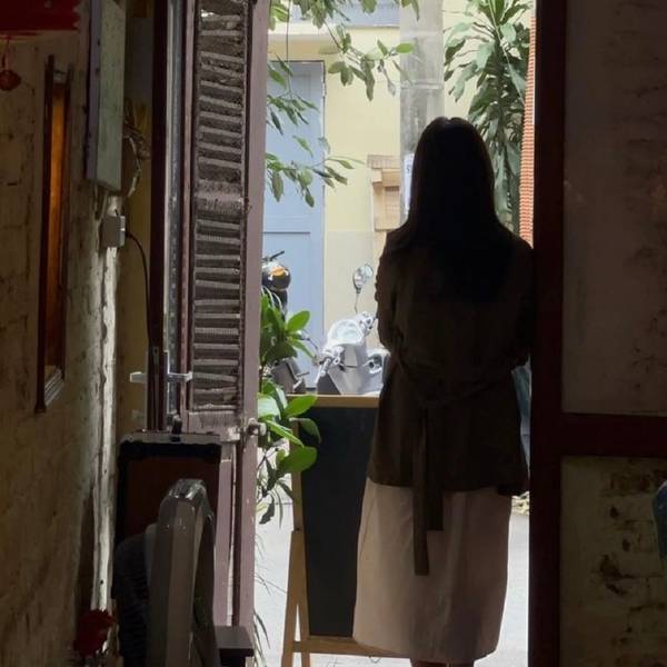 Người đứng quay lưng trong khung cửa, nh&igrave;n ra con hẻm nhỏ c&oacute; xe m&aacute;y v&agrave; c&acirc;y xanh.