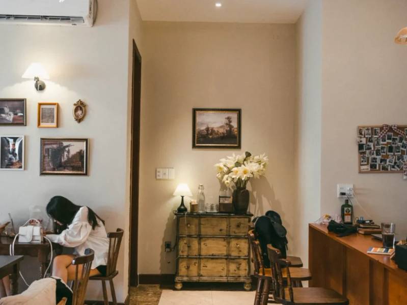 Qu&aacute;n c&agrave; ph&ecirc; với nội thất ấm c&uacute;ng, c&oacute; b&agrave;n ghế gỗ, tranh treo tường v&agrave; một người ngồi ở g&oacute;c đang sử dụng điện thoại.