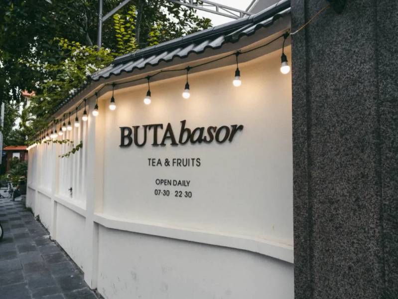 Bức tường trắng với đ&egrave;n trang tr&iacute; v&agrave; bảng hiệu qu&aacute;n tr&agrave;, tr&aacute;i c&acirc;y mở cửa h&agrave;ng ng&agrave;y từ 07:30 đến 22:30.