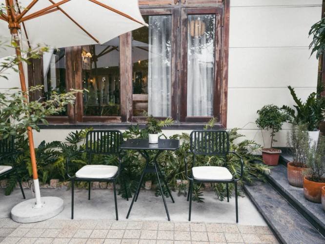 Bàn ghế ngoài trời với dù che, cây xanh, trước cửa sổ gỗ của quán cà phê.