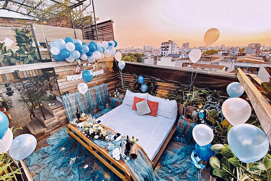 Giường trang tr&iacute; với b&oacute;ng bay xanh trắng, hoa v&agrave; gối, tr&ecirc;n s&acirc;n thượng nh&igrave;n ra th&agrave;nh phố l&uacute;c ho&agrave;ng h&ocirc;n.