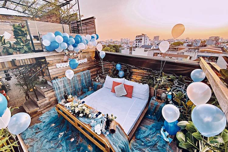 Sân thượng lãng mạn với giường trang trí và bóng bay xanh trắng, nhìn ra thành phố lúc hoàng hôn.