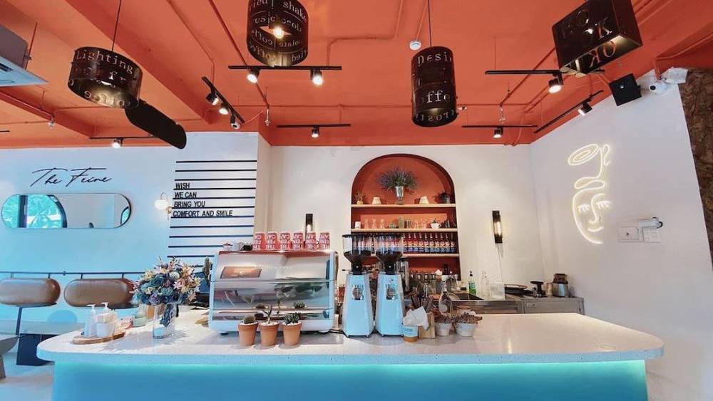 Qu&aacute;n c&agrave; ph&ecirc; với thiết kế hiện đại, b&agrave;n quầy bar trắng, đ&egrave;n trần, trang tr&iacute; hoa v&agrave; kệ trưng b&agrave;y sau quầy.