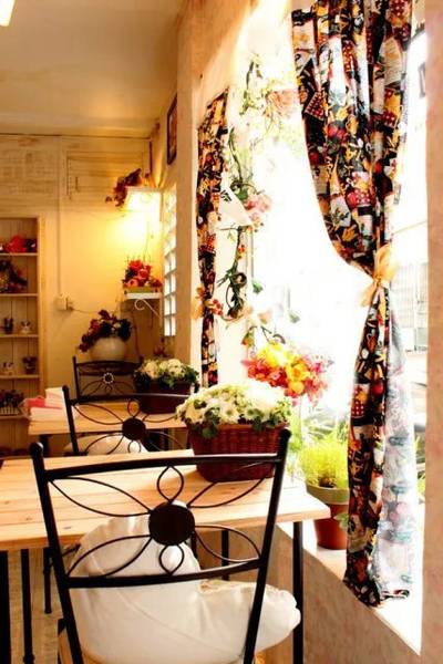 Qu&aacute;n c&agrave; ph&ecirc; nhỏ với b&agrave;n ghế gỗ, r&egrave;m cửa họa tiết, hoa trang tr&iacute; v&agrave; &aacute;nh s&aacute;ng tự nhi&ecirc;n từ cửa sổ.