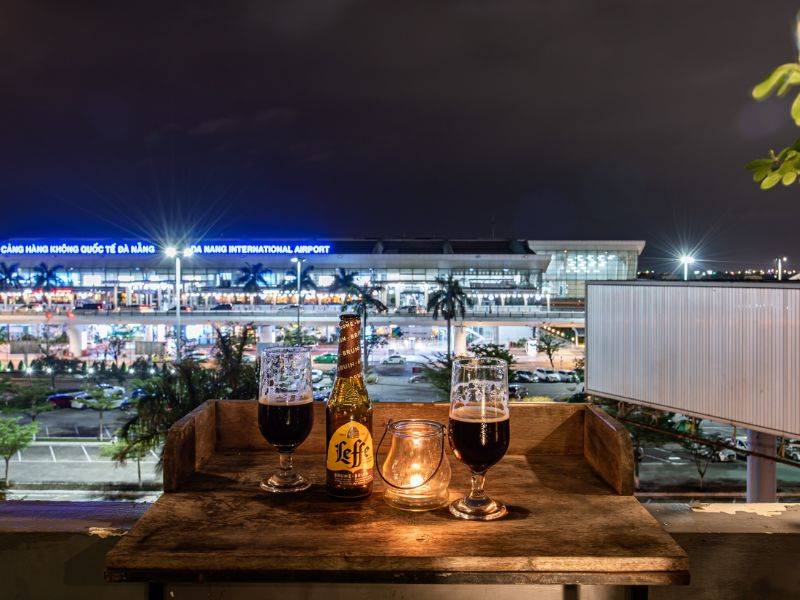 Hai ly bia v&agrave; một chai tr&ecirc;n b&agrave;n, nền l&agrave; s&acirc;n bay Đ&agrave; Nẵng v&agrave;o ban đ&ecirc;m với &aacute;nh s&aacute;ng rực rỡ.