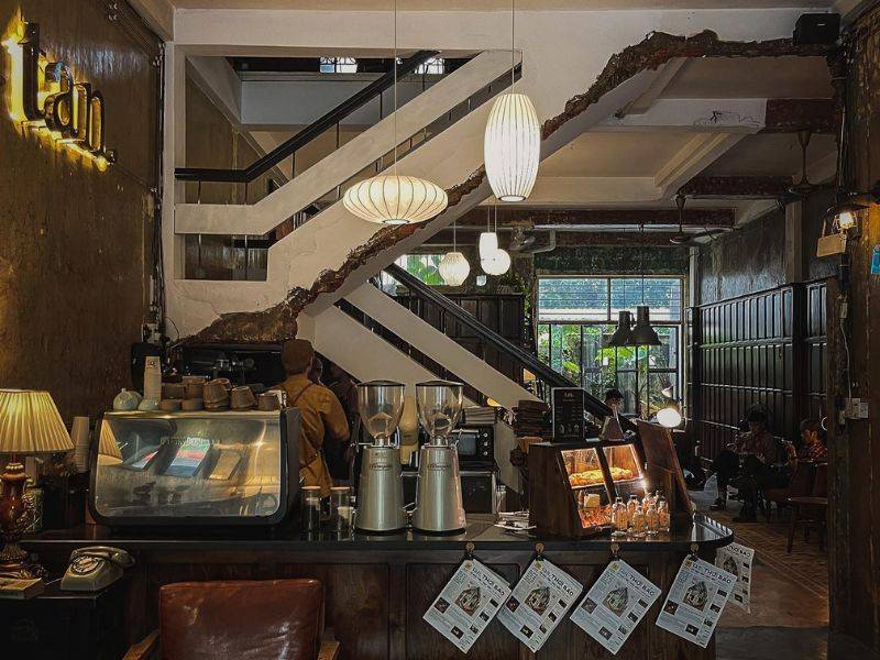 Qu&aacute;n c&agrave; ph&ecirc; với quầy pha chế, cầu thang, &aacute;nh s&aacute;ng dịu, v&agrave; kh&aacute;ch ngồi thư gi&atilde;n trong kh&ocirc;ng gian ấm c&uacute;ng.