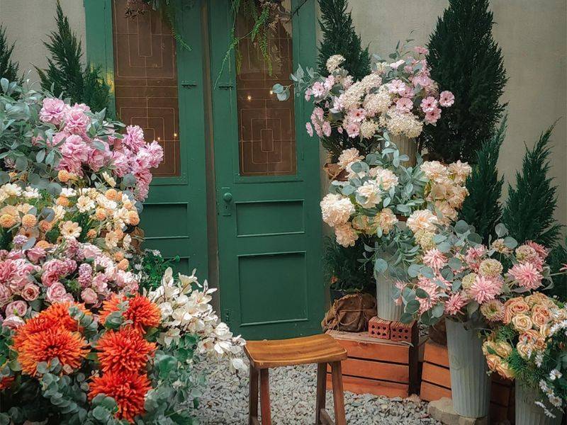 Cửa m&agrave;u xanh với c&aacute;c chậu hoa đa sắc xung quanh, ghế gỗ nhỏ đặt trước cửa, khung cảnh y&ecirc;n b&igrave;nh.