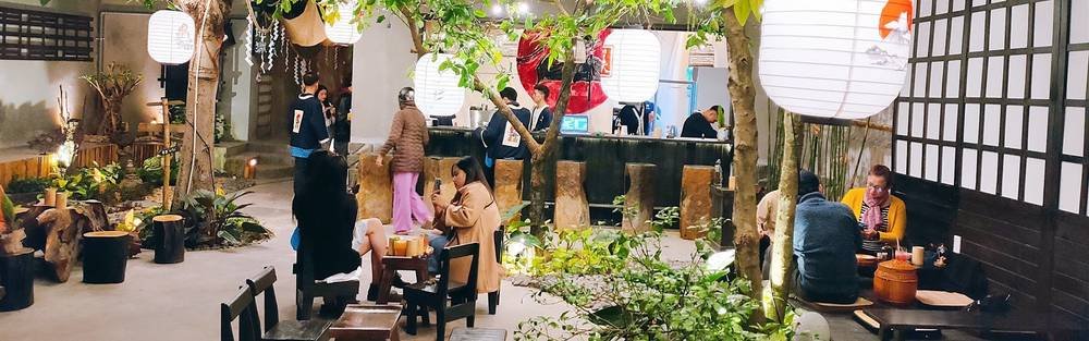 Quán cà phê phong cách Nhật Bản với lồng đèn sáng, cây xanh xung quanh và khách hàng trò chuyện vui vẻ.