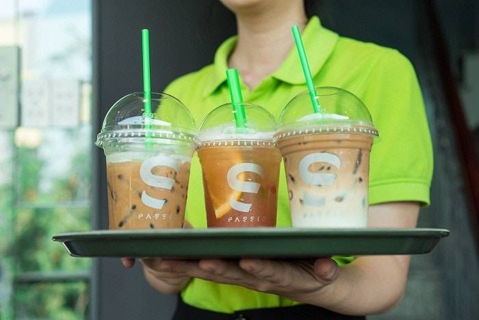 Người cầm khay với ba ly đồ uống c&oacute; ống h&uacute;t m&agrave;u xanh, mặc &aacute;o xanh l&aacute;, nền mờ.