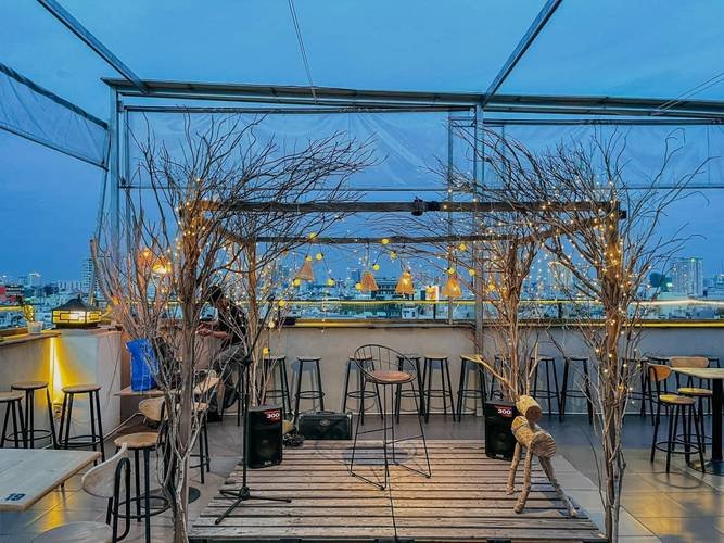Sân thượng quán cà phê với ánh đèn vàng lung linh, ghế ngồi thoải mái, cây trang trí và tầm nhìn thành phố ban đêm.
