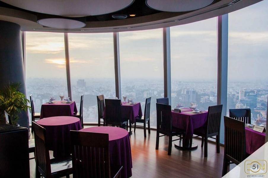 Nh&agrave; h&agrave;ng tr&ecirc;n cao với b&agrave;n ghế gỗ, khăn trải b&agrave;n t&iacute;m, cửa sổ lớn nh&igrave;n ra khung cảnh th&agrave;nh phố l&uacute;c ho&agrave;ng h&ocirc;n.