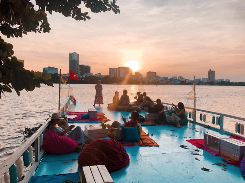 Một nh&oacute;m người ngồi thư gi&atilde;n tr&ecirc;n thuyền, ngắm ho&agrave;ng h&ocirc;n tr&ecirc;n s&ocirc;ng, với bối cảnh th&agrave;nh phố ph&iacute;a xa.