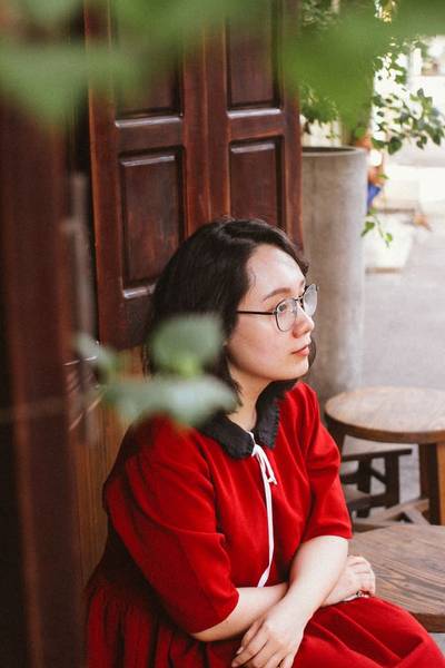 Người phụ nữ mặc &aacute;o đỏ ngồi b&ecirc;n cửa gỗ, nh&igrave;n ra xa với vẻ trầm tư, xung quanh l&agrave; c&acirc;y xanh v&agrave; ghế gỗ.