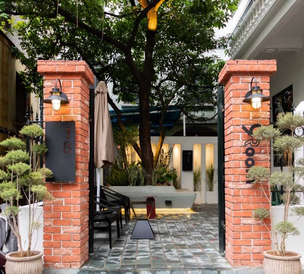 Khám Phá Top Quán Cafe Đẹp Ở Liễu Giai: Đồ Uống Ngon, Không Gian Thư Giãn 17 Cổng gạch đỏ dẫn vào khu vườn nhỏ với nhiều cây xanh và ghế ngồi, có đèn chiếu sáng và số nhà.