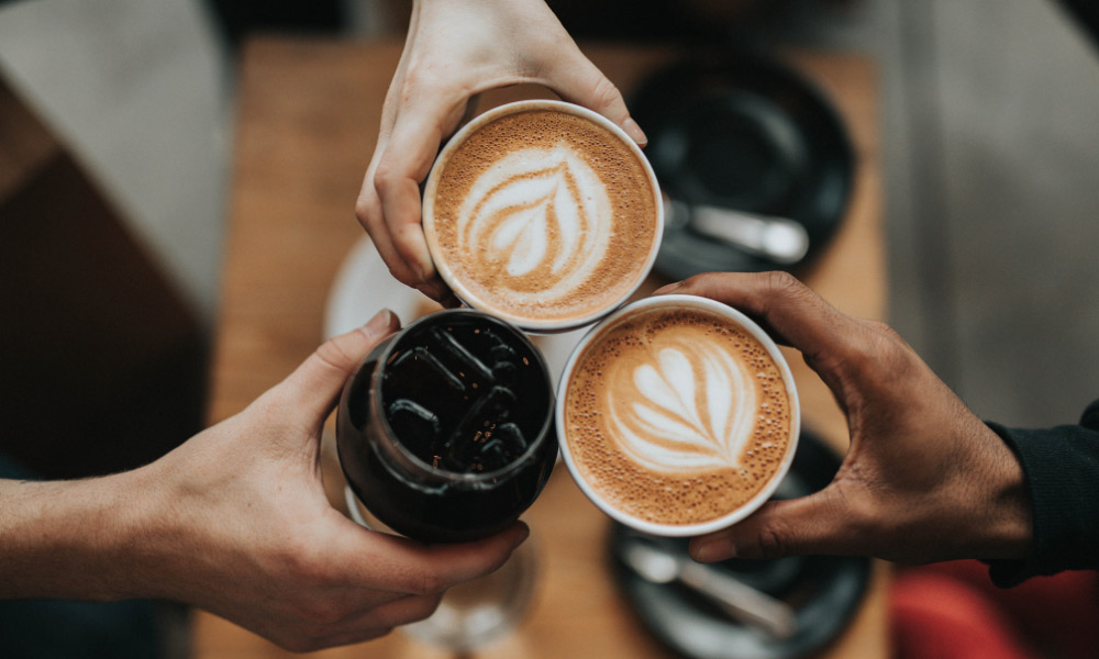 Khám Phá 14 Quán Cafe Hồ Tùng Mậu: Điểm Chill Lý Tưởng Tại Cầu Giấy 21 Ba người cầm ly cà phê và đồ uống lạnh, đang cụng ly trên bàn gỗ.
