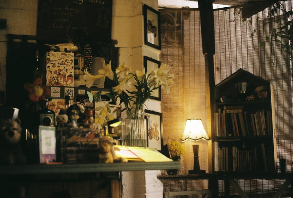 Khám Phá 15+ Quán Cafe View Đẹp, Đồ Uống Ngon Tại Hoàng Cầu 6 Góc phòng với kệ sách, đèn bàn sáng nhẹ, bình hoa trang trí và tranh ảnh trên tường, tạo không gian ấm cúng.