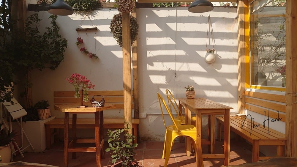15 Quán Cafe Chùa Láng Với Không Gian Thư Giãn Tuyệt Đẹp 20 Quán cà phê ngoài trời với bàn ghế gỗ, ghế kim loại vàng, cây xanh và ánh sáng tự nhiên chiếu qua giàn che.
