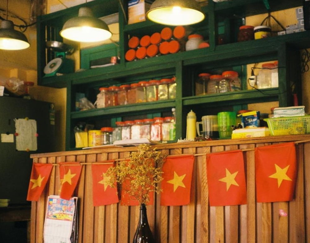 Khám Phá 15+ Quán Cafe View Đẹp, Đồ Uống Ngon Tại Hoàng Cầu 21 Quầy gỗ trang trí cờ đỏ sao vàng, trước kệ chứa lọ thủy tinh và vật dụng, chiếu sáng bởi đèn treo.