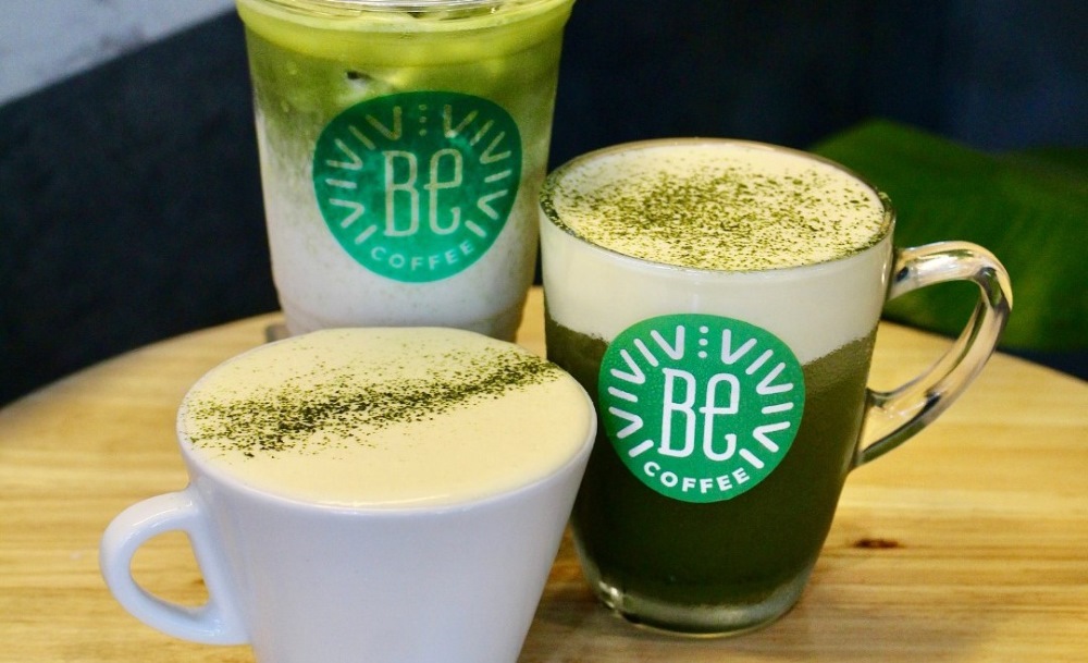 Khám Phá Top 10 Quán Cafe Giảng Võ Hấp Dẫn Nhất Tại Hà Nội 14 Ba ly đồ uống với bọt sữa và bột trà xanh, đặt trên bàn gỗ, có logo BE Coffee màu xanh.