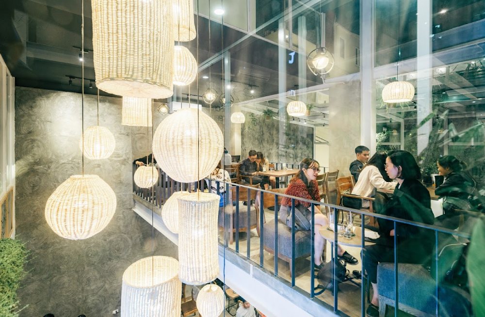 Khám Phá 7 Quán Cafe Tuyệt Đẹp Ở Ngã Tư Sở: View Đẹp, Đồ Uống Ngon, Giá Phải Chăng 10 Quán cà phê với thiết kế hiện đại, ánh sáng ấm áp từ đèn treo, khách ngồi trò chuyện bên bàn gỗ.