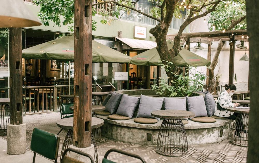 Khám Phá 7 Quán Cafe Tuyệt Đẹp Ở Ngã Tư Sở: View Đẹp, Đồ Uống Ngon, Giá Phải Chăng 6 Quán cà phê ngoài trời với cây xanh, dù che nắng, bàn ghế sắp xếp xung quanh và một người đang ngồi.