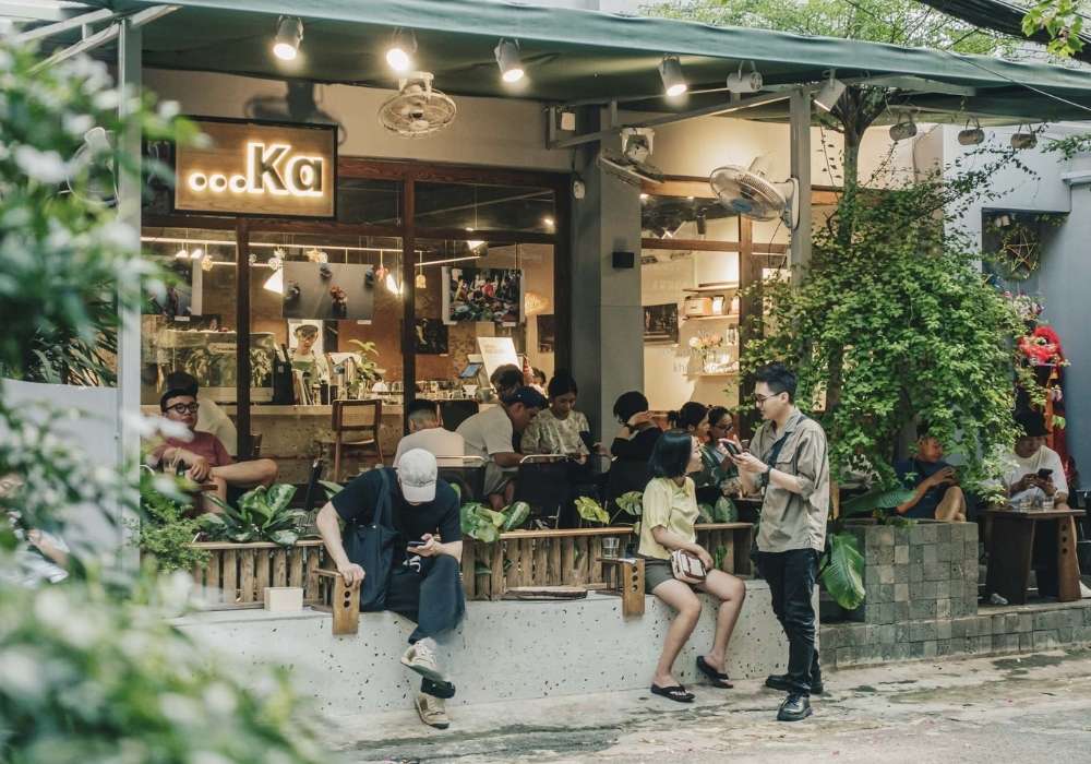 Khám Phá Top 10 Quán Cafe Láng Hạ: View Đẹp, Menu Hấp Dẫn, Giá Cực Hợp Lý 19 Quán cà phê đông đúc với nhiều người ngồi trò chuyện và sử dụng điện thoại, cây xanh bao quanh.