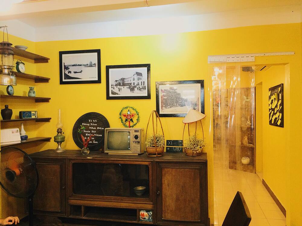 Khám Phá 10+ Quán Cafe Đẹp Ở Đặng Văn Ngữ: Đồ Uống Ngon, Giá Cả Hấp Dẫn 26 Phòng khách với tường vàng, tivi cổ, tranh đen trắng, kệ để đồ và quạt bàn trong không gian trang trí hoài cổ.