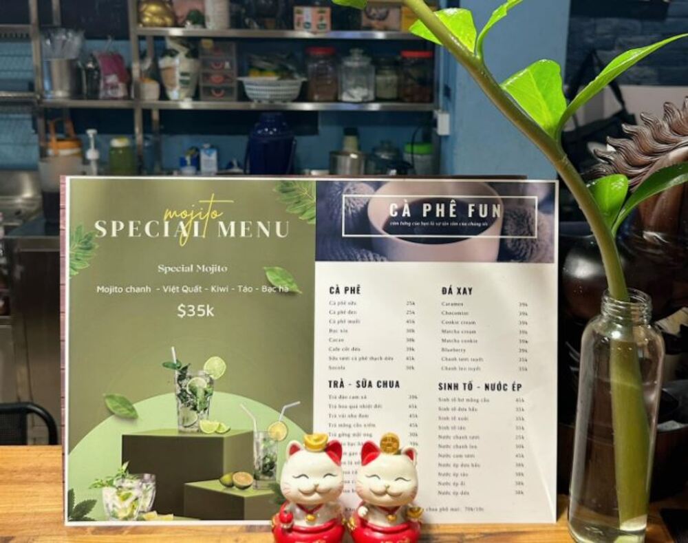 Khám Phá 10+ Quán Cafe Đẹp Ở Đặng Văn Ngữ: Đồ Uống Ngon, Giá Cả Hấp Dẫn 8 Thực đơn cà phê và mojito đặt trên quầy, bên cạnh có hai tượng mèo và một bình cắm cây xanh.