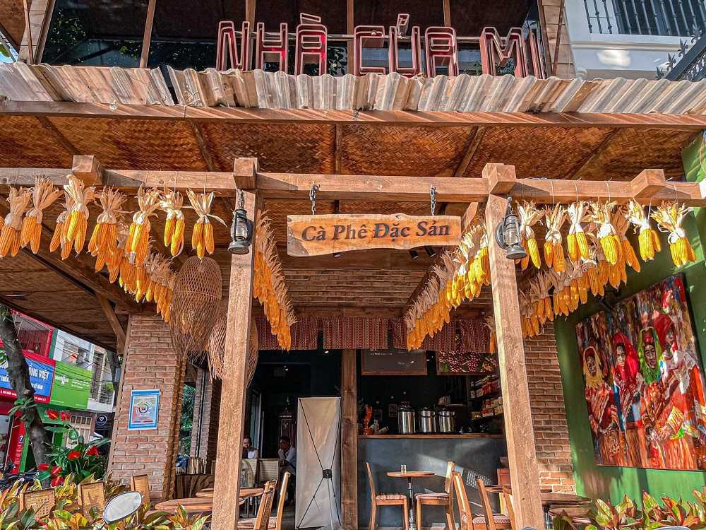 Khám Phá Những Quán Cafe Độc Lạ Thu Hút Giới Trẻ Tại Sài Gòn 17 Quán cà phê với bảng hiệu "Cà Phê Đặc Sản", trang trí ngô treo, không gian gỗ, có tranh treo tường.