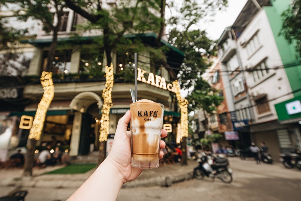 Khám Phá 15+ Quán Cafe View Đẹp, Đồ Uống Ngon Tại Hoàng Cầu 14 Người cầm ly cà phê trước quán cà phê có đèn trang trí, nằm trên con phố với nhiều xe máy và tòa nhà.