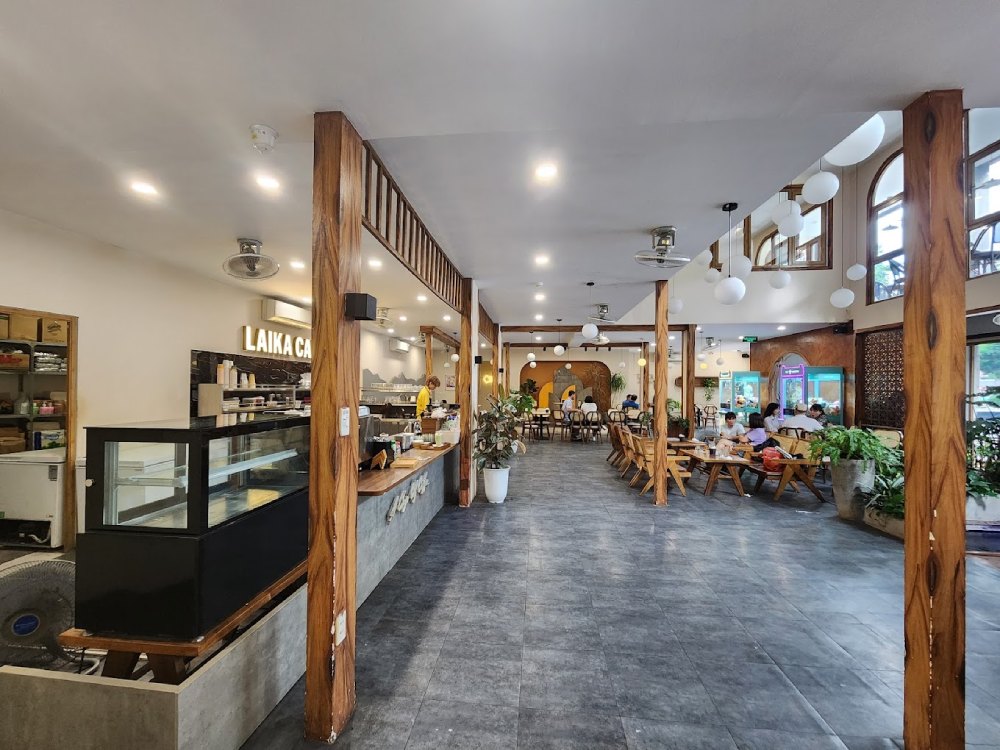 Khám Phá 7 Quán Cafe Tuyệt Đẹp Ở Ngã Tư Sở: View Đẹp, Đồ Uống Ngon, Giá Phải Chăng 12 Quán cà phê rộng rãi với bàn ghế gỗ, cây xanh trang trí và quầy phục vụ, khách đang ngồi thư giãn.