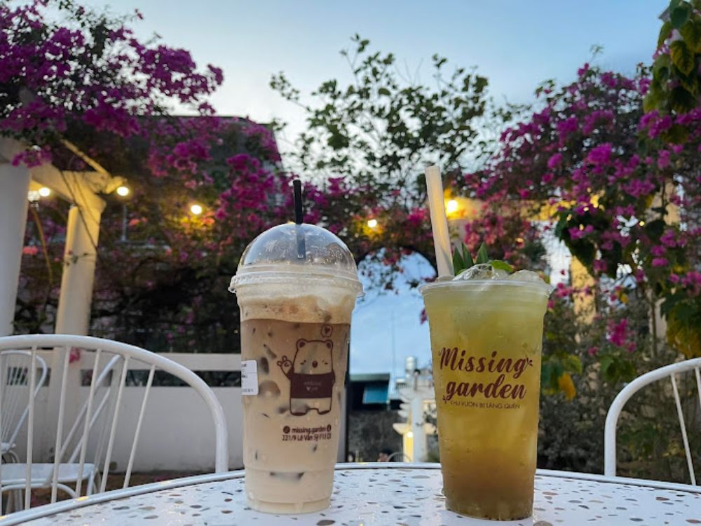 Khám Phá Top 6 Quán Cafe Mở Cửa 24h Tại Quận 3 Cho Người Yêu Thích Cú Đêm 5 Hai ly đồ uống trên bàn ngoài trời, nền là hoa giấy tím và bầu trời xanh.